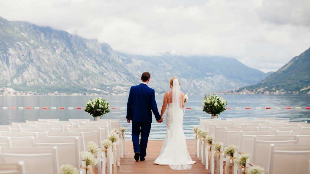 Man and woman holding hands and walking down a dock with chairs and flowers.